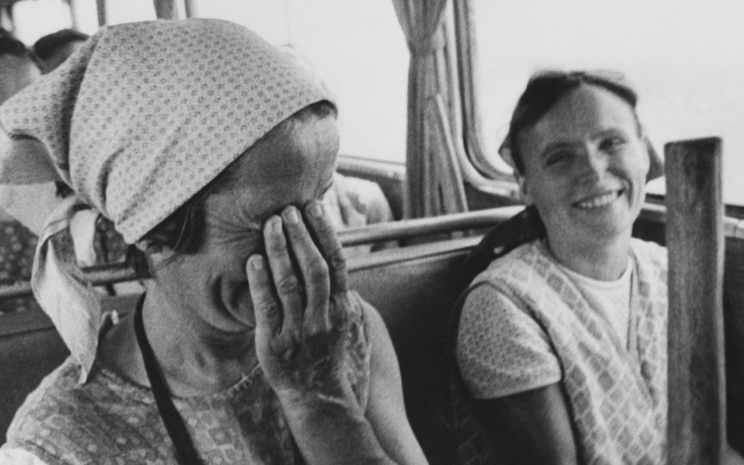 Filmstill aus ISTENMEZEJE, EIN UNGARISCHES DORF: Zwei Frauen in bäuerlicher Kleidung sitzen in einem Bus. Beide lachen, eine hält sich die Hand vors Gesicht.