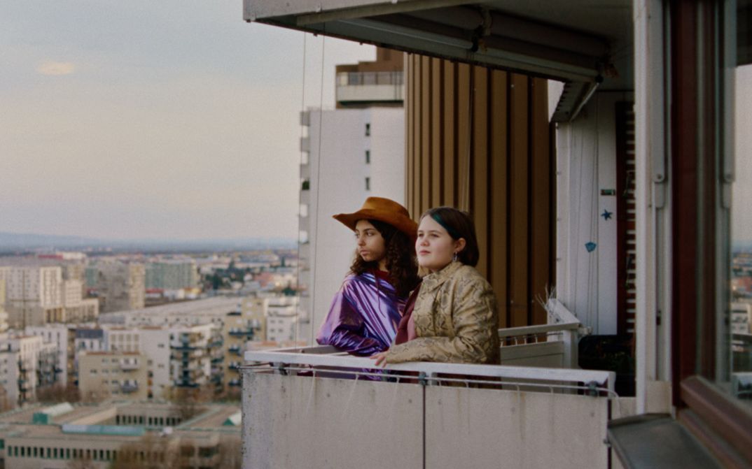 Film still from WENN DU ANGST HAST NIMMST DU DEIN DEIN HERZ IN DEN MUND UND LÄCHELST by Marie-Luise Lehner. Two young women stand on the balcony of a high-rise building and look into the distance.