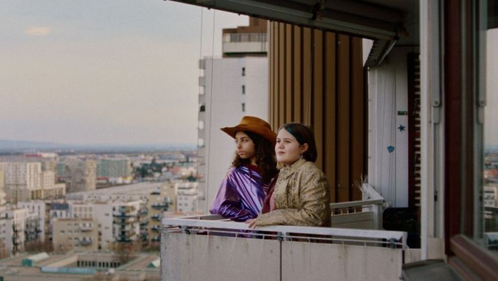Film still from WENN DU ANGST HAST NIMMST DU DEIN DEIN HERZ IN DEN MUND UND LÄCHELST by Marie-Luise Lehner. Two young women stand on the balcony of a high-rise building and look into the distance. 