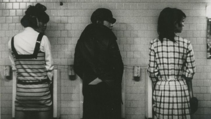 Film still from FUNERAL PARADE OF ROSES: Three people in extravagant dresses in front of three urinals, all looking to the right.