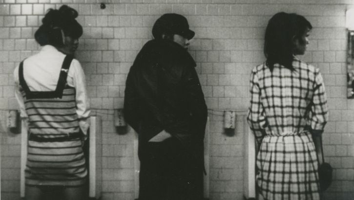 Filmstill aus FUNERAL PARADE OF ROSES: Drei Menschen in extravaganter Kleider vor drei Pissoirs, alle blicken nach rechts.
