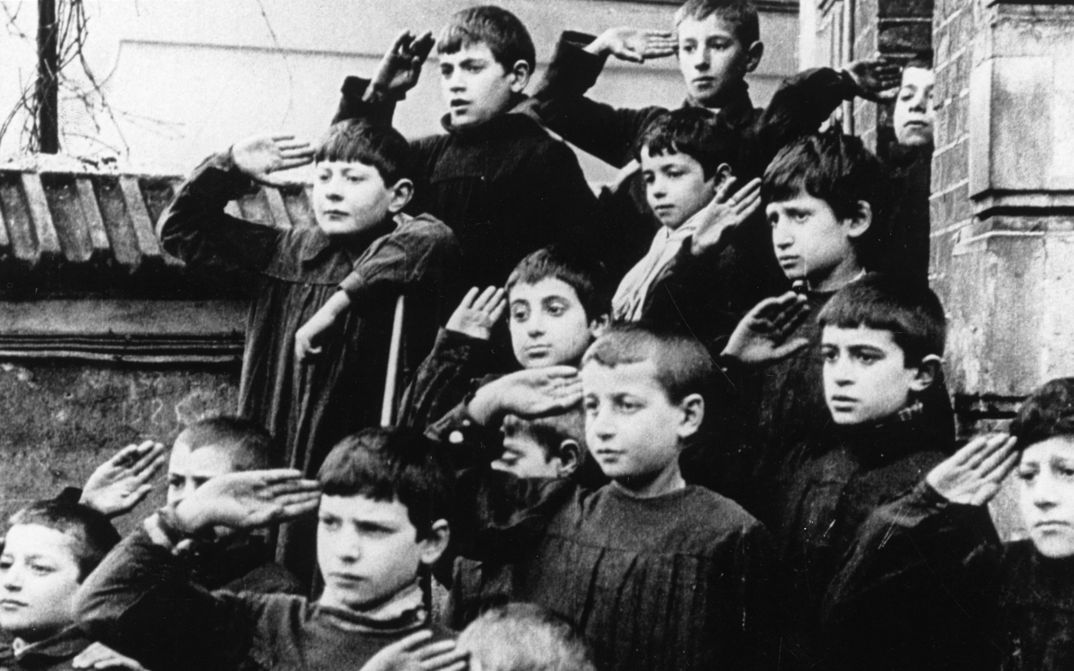 Filmstill aus ZÉRO DE CONDUITE: Eine Gruppe von Kindern vor einem Gebäude. Alle halten ihre rechte Hand an die Schläfe.