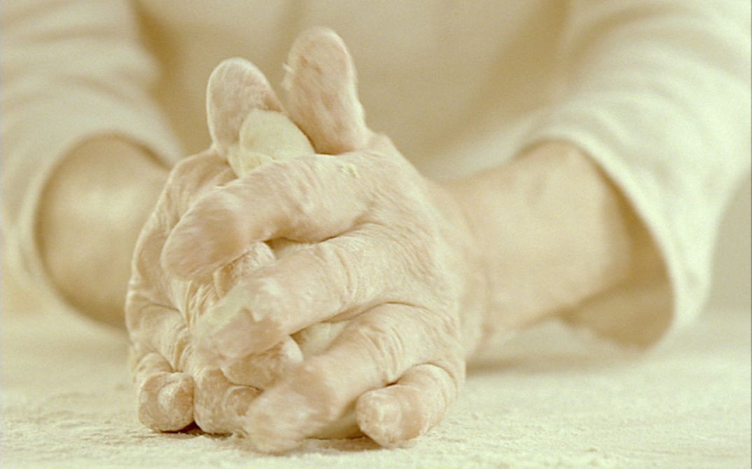 Filmstill aus CUM PANE. Großaufnahme von zwei Händen, die Teig kneten.