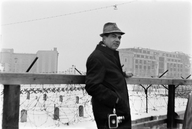 Film still from "Il cassetto segreto" by Costanza Quatriglio. It shows a black and white image of a man standing at a high-security fence. He is wearing a coat and a hat.