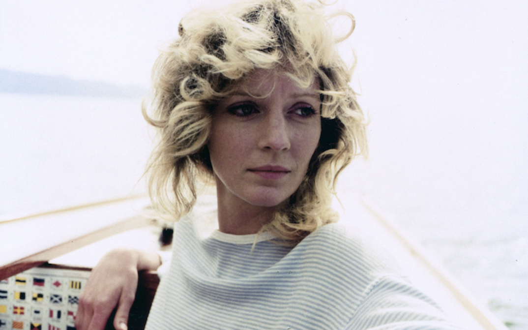 Film still from DIE ALLEINSEGLERIN: A young woman with curly blonde hair stands at the bow of a sailing boat.