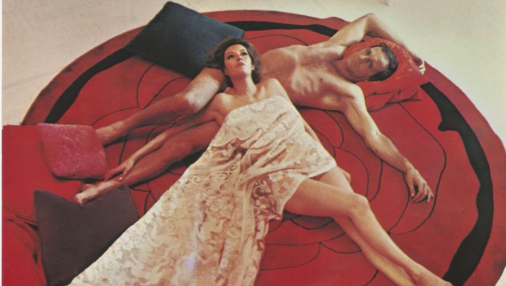 Film still from COME, QUANDO, PERCHÉ: A man and a woman lie naked on a round red carpet. Her body is half covered by a blanket.