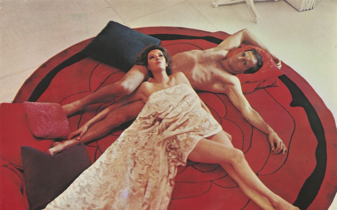 Film still from COME, QUANDO, PERCHÉ: A man and a woman lie naked on a round red carpet. Her body is half covered by a blanket.