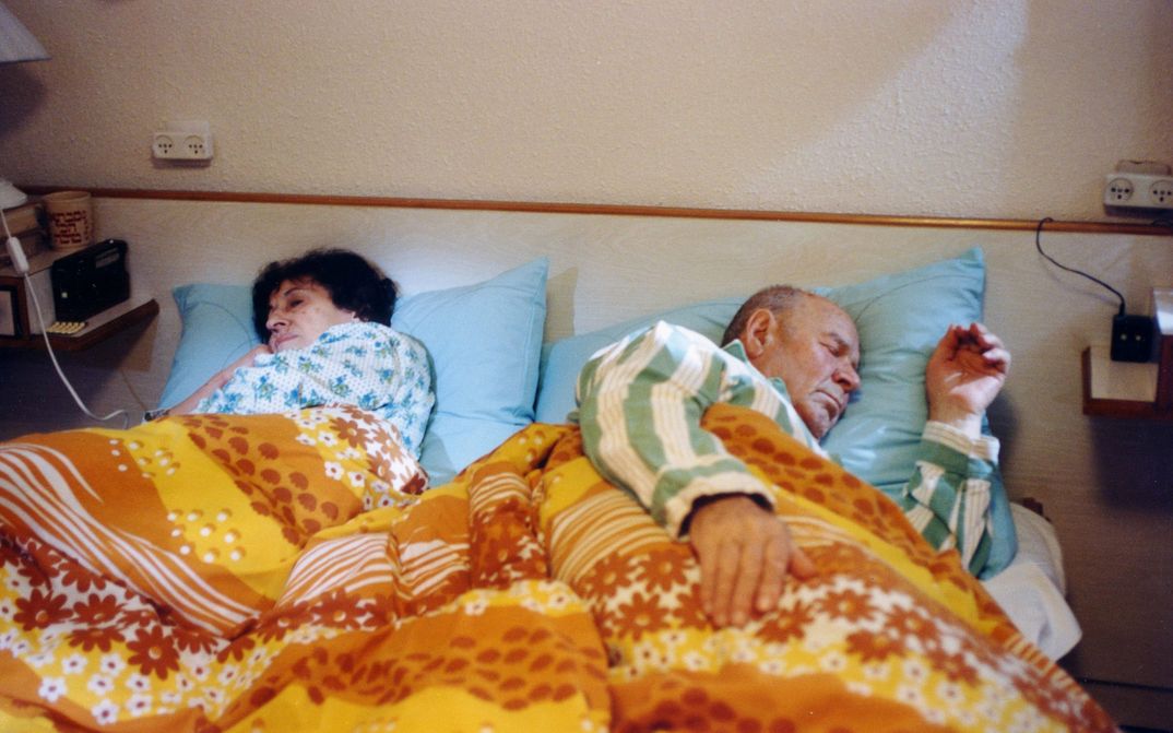 Filmstill from HABEHIRA VEHAGORAL: A woman and a man lie asleep in bed. The bed linen and pyjamas are patterned and colourful.
