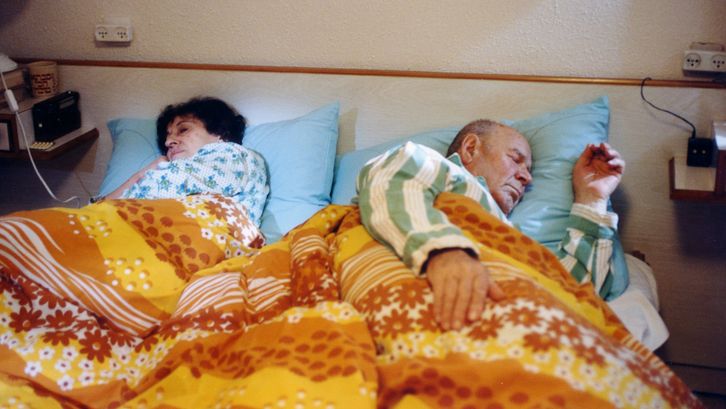 Filmstill aus HABEHIRA VEHAGORAL: Eine Frau und ein Mann liegen schlafend im Bett. Die Bettwäsche sowie Schlafanzüge sind gemustert und farbenfroh.