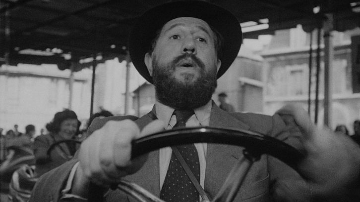 Film still from PANIQUE: A man in a suit and hat rides a bumper car alone.