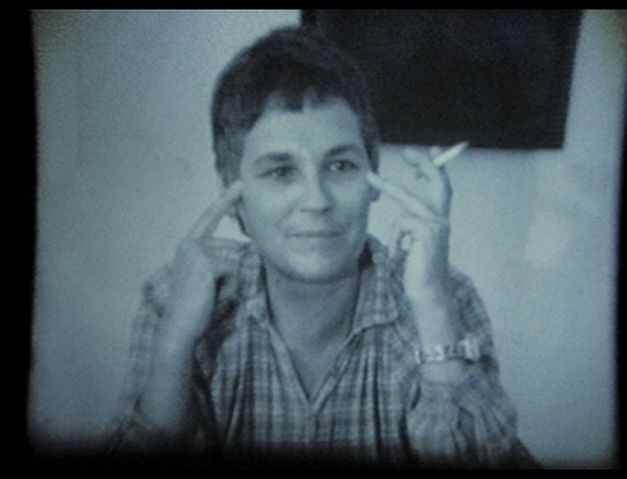 A woman sits at a table, points to her temples with her fingers and holds a cigarette.