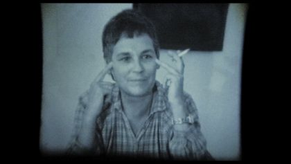 Frau sitzt an einem Tisch, zeigt mit den Fingern auf ihre Schläfen und hält eine Zigarette.