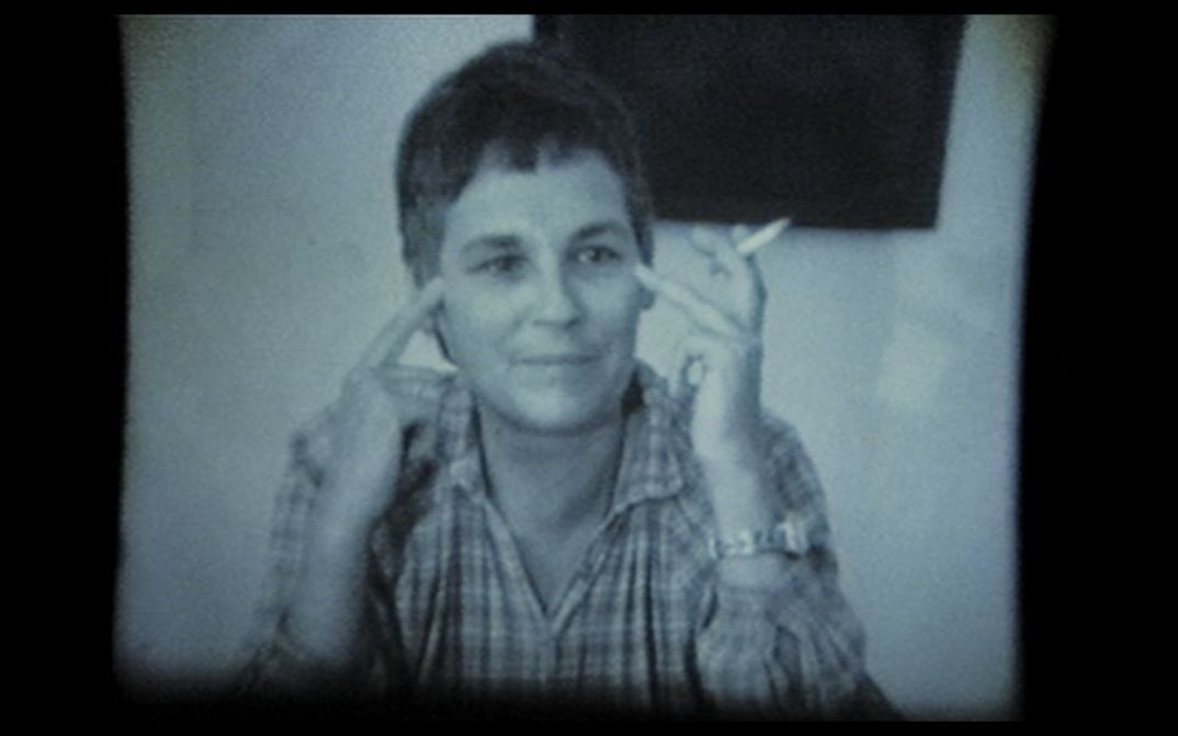 Frau sitzt an einem Tisch, zeigt mit den Fingern auf ihre Schläfen und hält eine Zigarette.