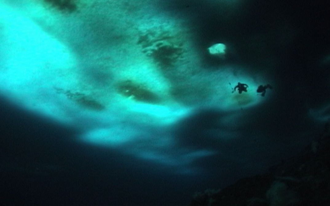 Film still from THE WILD BLUE YONDER: An underwater landscape in dark blue and turquoise. In the background you can see two people diving.
