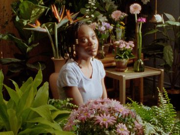 Filmstill aus „This House“ von Miryam Charles.Ein Mädchen sitzt auf dem Boden eines hellen Zimmers und schaut hoch. Um sie herum viele Blumen.