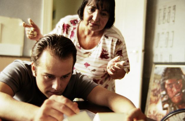 Still from the film "FREMD. YABAN." by Hakan Savaş Mican. A young man leans over a table while an older woman watches over his shoulder. 