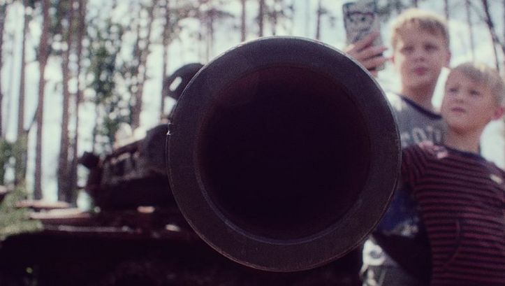 An armoured flexible tube is pointed at the camera, while in the background two children take a picture of it with a cell phone.