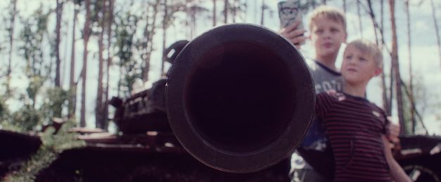 An armoured flexible tube is pointed at the camera, while in the background two children take a picture of it with a cell phone.