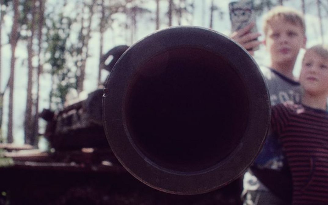 An armoured flexible tube is pointed at the camera, while in the background two children take a picture of it with a cell phone.