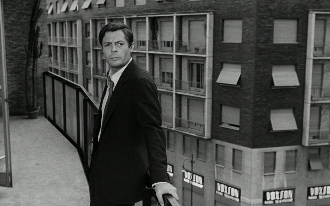 Filmstill aus LA NOTTE: Ein Mann steht an einem Balkongeländer, hinter ihm ist die Fassade eines Hauses.