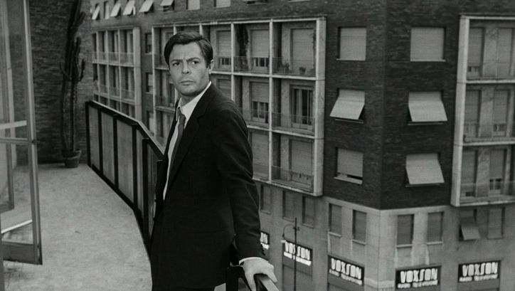 Film still from LA NOTTE: A man stands at a balcony railing, behind him is the facade of a house.