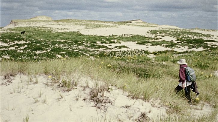Filmstill aus „Geographies of Solitude“ von Jacquelyn Mills. In einer Dünenlandschaft in der Sonne läuft eine Frau. Sie trägt eine dunkelrote Windjacke, einen Rucksack und einen Fischerhut.