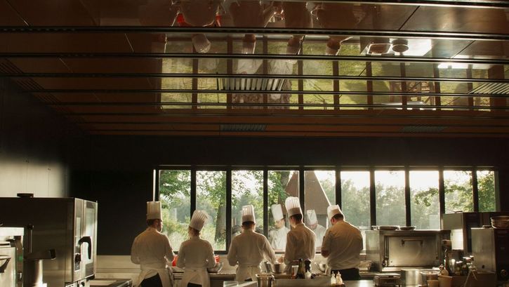Film still from MENUS PLAISIRS: Five chefs stand next to each other in the kitchen. They can be seen twice through a reflection.