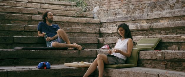 A man and a woman are sitting outside on wooden steps with cushions. Next to them is a plate of food and a glass of water. A pink roller skate lies between the woman and the man. 