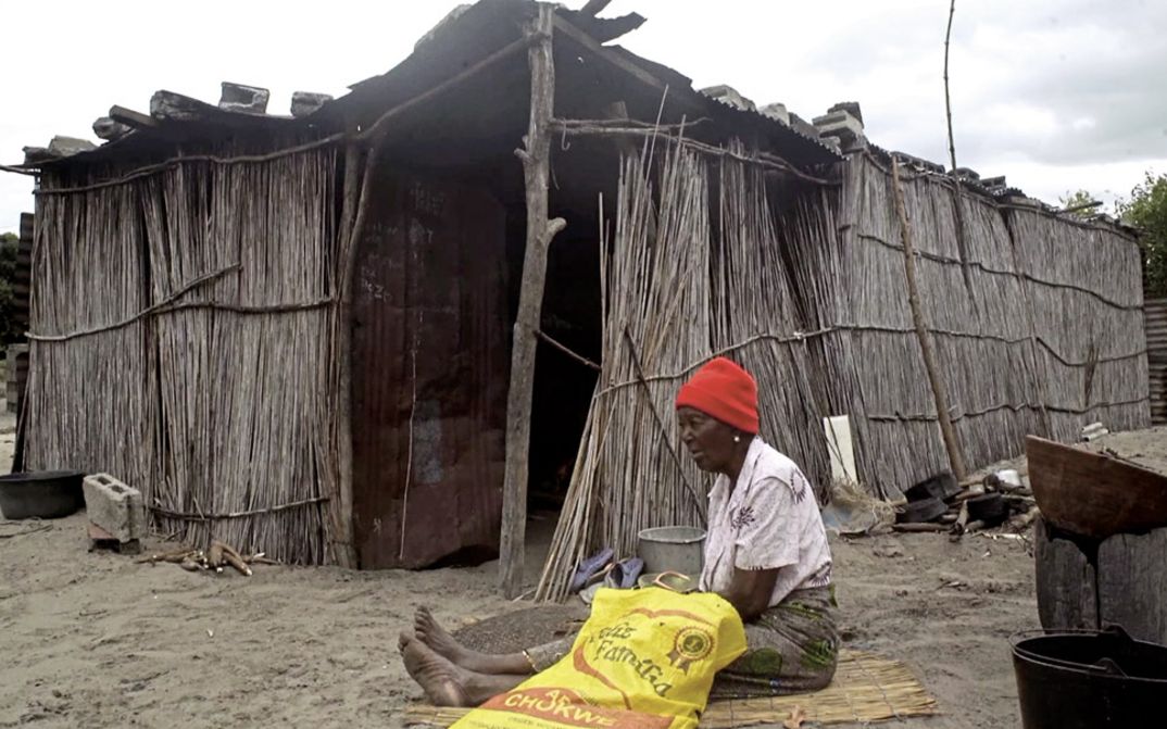 Film still from "The Nights Still Smell of Gunpowder" by Inadelso Cossa. A woman with a red beanie is sitting on the ground in front of a cabin made of sticks.