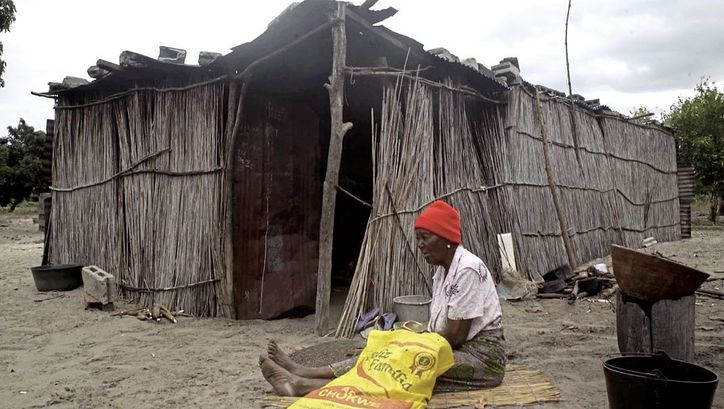 Film still from "The Nights Still Smell of Gunpowder" by Inadelso Cossa. A woman with a red beanie is sitting on the ground in front of a cabin made of sticks.