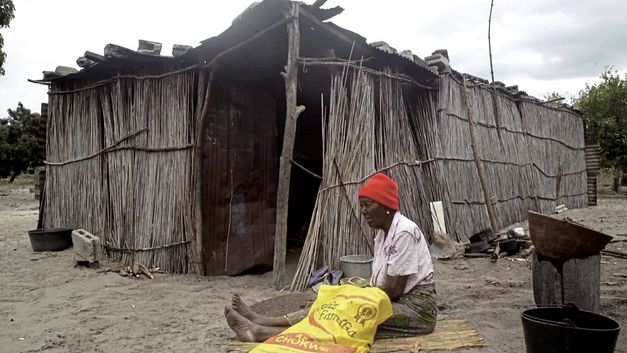 Film still from "The Nights Still Smell of Gunpowder" by Inadelso Cossa. A woman with a red beanie is sitting on the ground in front of a cabin made of sticks.