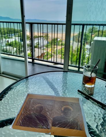 Still from the making of "Parasite Family" by Prapat Jiwarangsan. Painted portrait photos lie on a glass table next to a drink. In the background you can see a balcony, the sea and the blue sky.