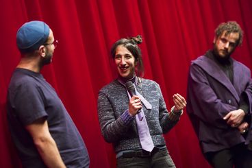 Three people in front of a red curtain with microphones are speaking to each other