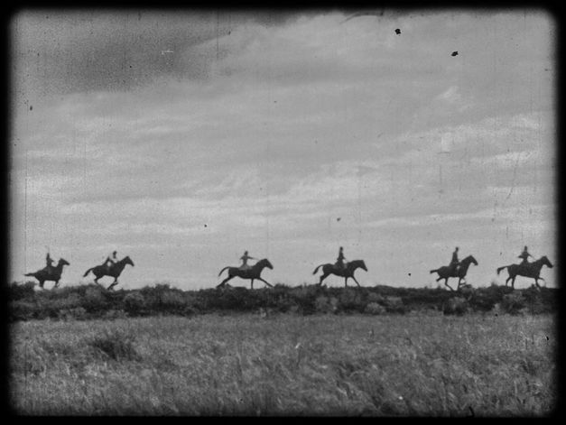 It shows a black and white picture of a meadow landscape. Six people on horseback ride one after the other through the landscape.