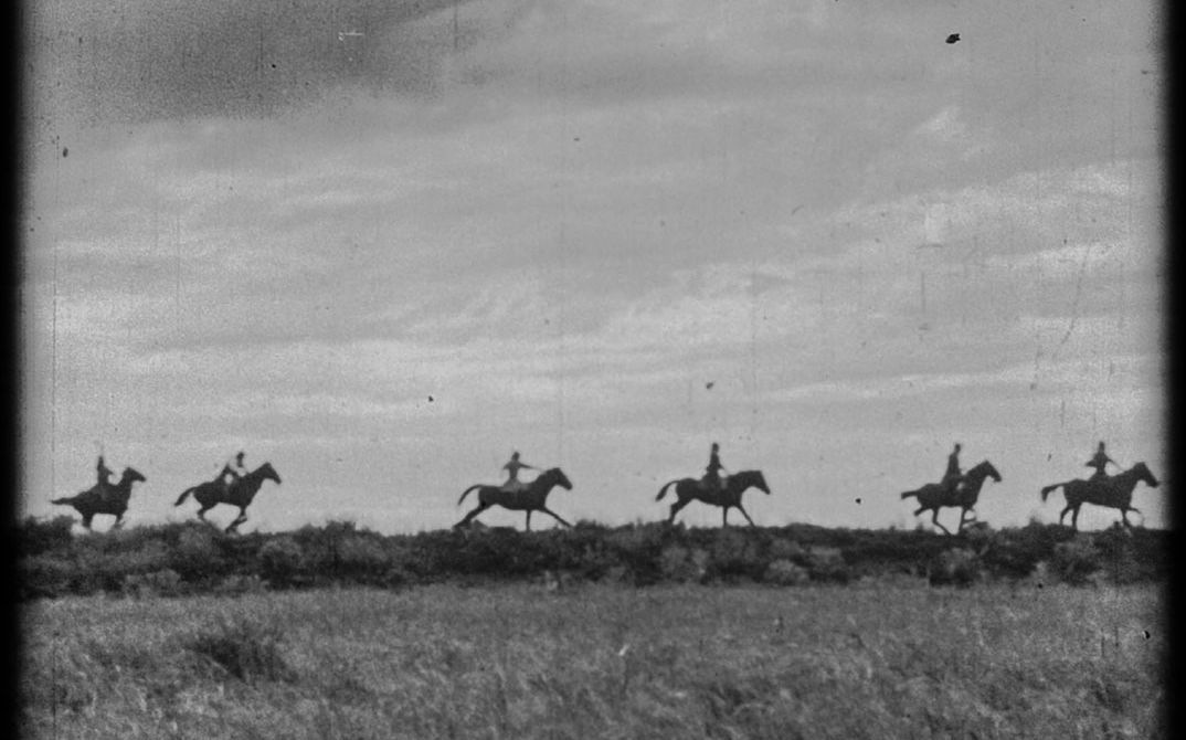 It shows a black and white picture of a meadow landscape. Six people on horseback ride one after the other through the landscape.