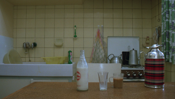 Filmstill aus JEANNE DIELMAN: Man sieht eine Küche, im Hintergrund Spülbecken und Herd, im Vordergrun ein Tisch, auf dem eine Milchflasche und Gläser stehen.