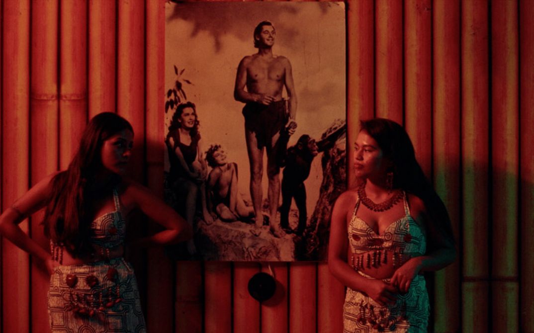 Two women stand in front of a wall of bamboo poles on which hangs a picture of Johnny Weissmuller as Tarzan.