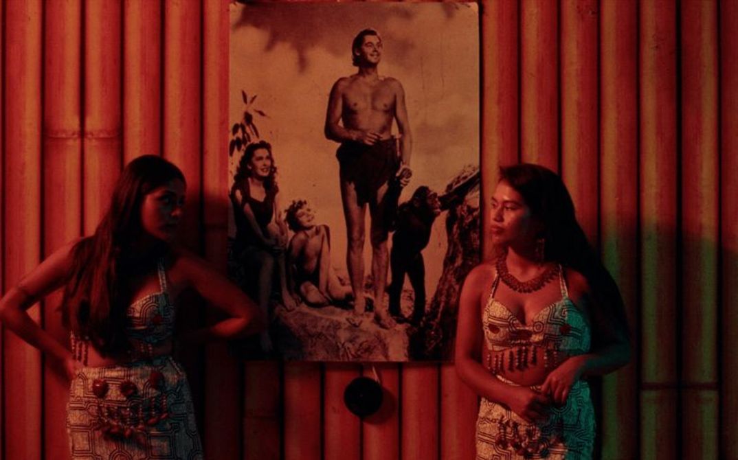 Two women stand in front of a wall of bamboo poles on which hangs a picture of Johnny Weissmuller as Tarzan.