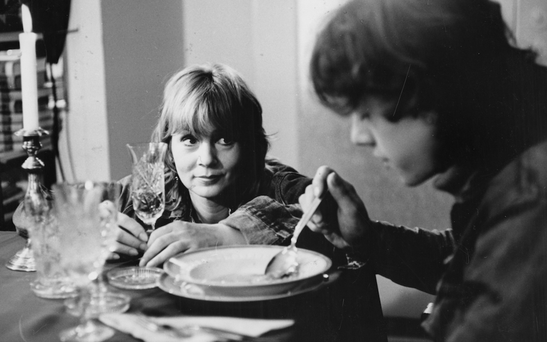 Film still from DIE BEUNRUHIGUNG: A woman sits at the table and looks at her son, who is eating soup. 