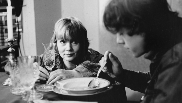 Film still from DIE BEUNRUHIGUNG: A woman sits at the table and looks at her son, who is eating soup. 