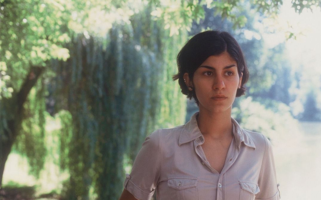 Filmstill aus DER SCHÖNE TAG: Eine Frau mit kinnlangen Haaren schaut nach rechts. Im Hintergrund sind Bäume. Die Sonne scheint.