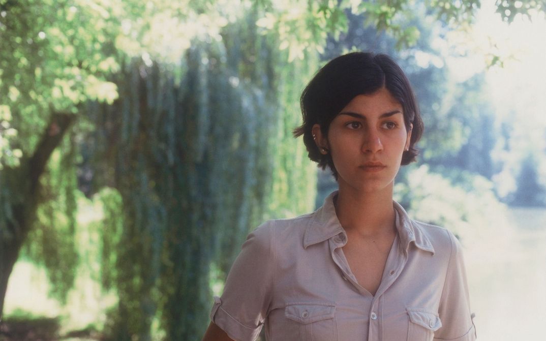 Film still from DER SCHÖNE TAG: A woman with chin-length hair looks to the right. There are trees in the background. The sun is shining.
