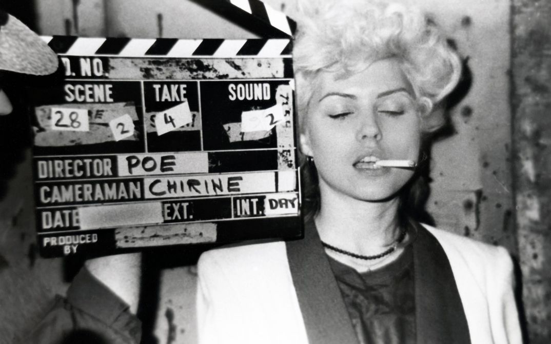 Filmstill from FOREIGNER: A woman with short blonde hair, a cigarette in the corner of her mouth and a white jacket has her eyes closed. A film clapperboard next to her.