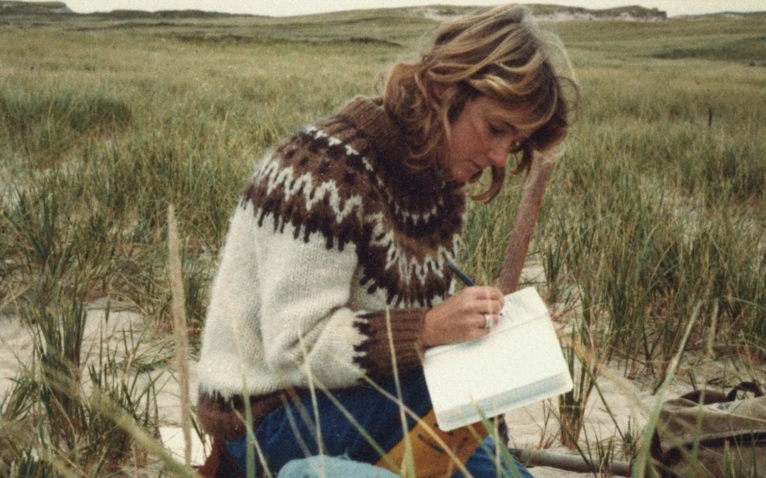 Filmstill aus „Geographies of Solitude“ von Jacquelyn Mills. Die Protagonistin Zoe Lucas sitzt mit Notizbuch in einer rauen Dünenlandschaft.