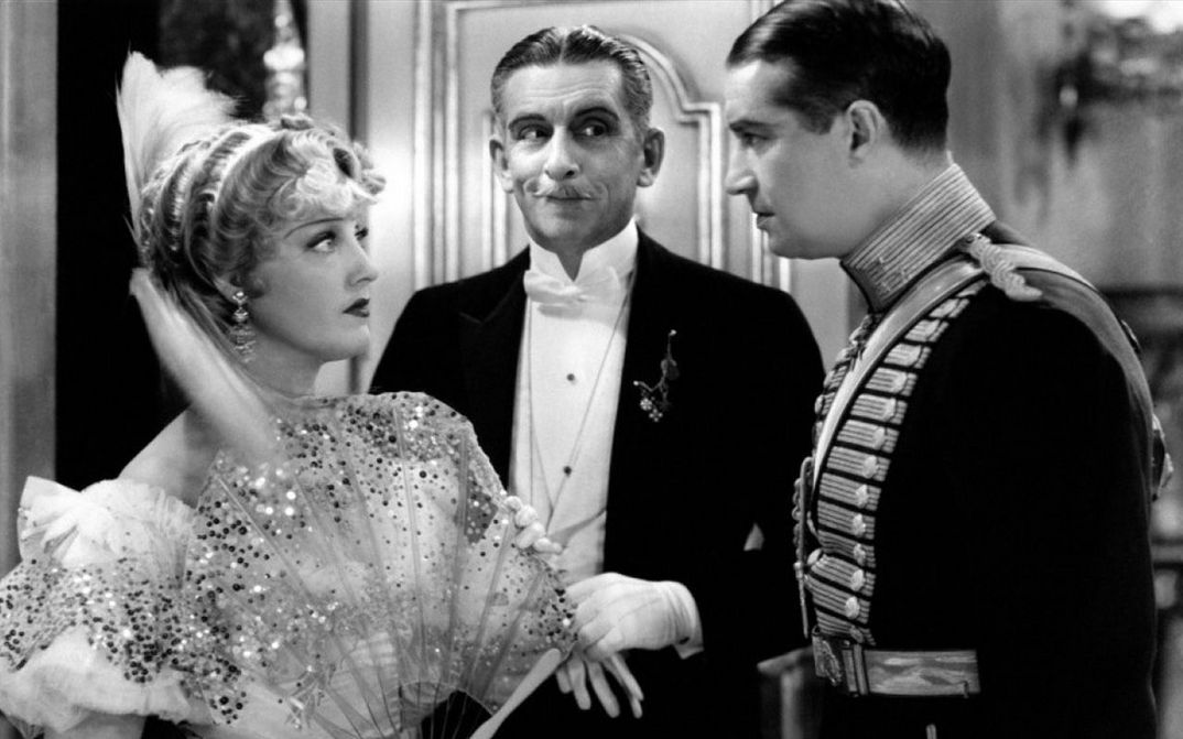 Filmstill aus THE MERRY WIDOW: Eine junge Frau und ein junger Mann, beide festlich gekleidet, blicken sich an. Im Hintergrund steht ein älterer Herr.
