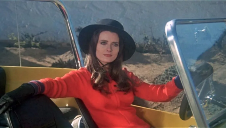 Film still from THE VELVET VAMPIRE: A young woman in a red dress, wearing a hat and gloves, sits at the wheel of an open car.