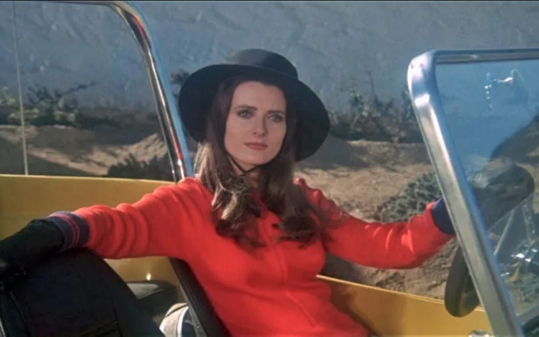 Film still from THE VELVET VAMPIRE: A young woman in a red dress, wearing a hat and gloves, sits at the wheel of an open car.