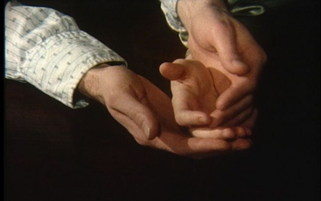 Filmstill aus NOW: Vor einem schwarzen Hintergrund sind man zwei Hände, die eine Kinderhand umschließen.