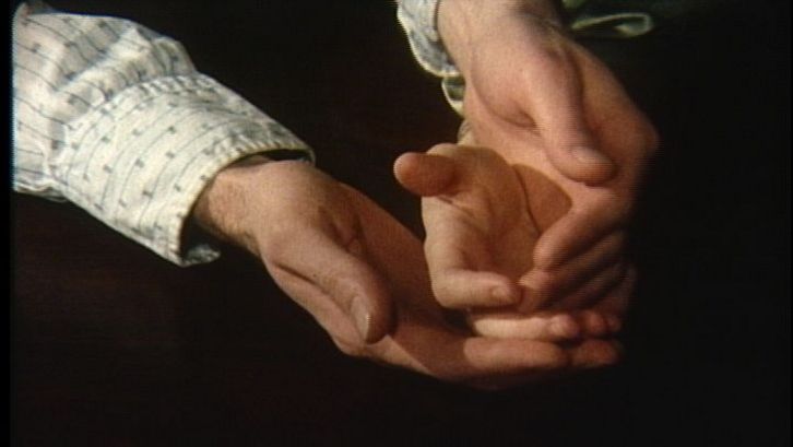Filmstill aus NOW: Vor einem schwarzen Hintergrund sind man zwei Hände, die eine Kinderhand umschließen.