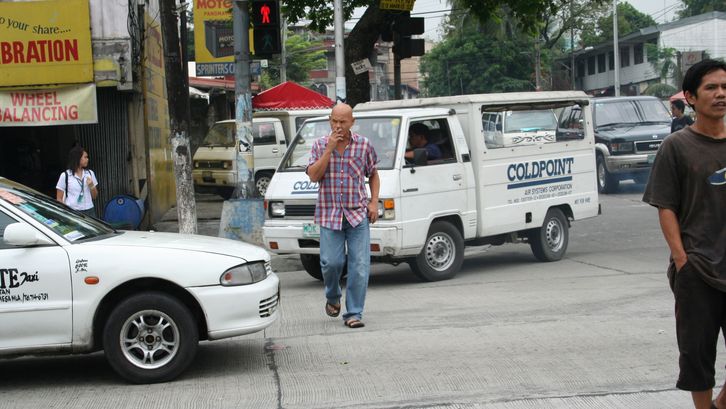 Film still from MANILA: A man crosses a road on which several cars are driving. He has a cigarette in his mouth.
